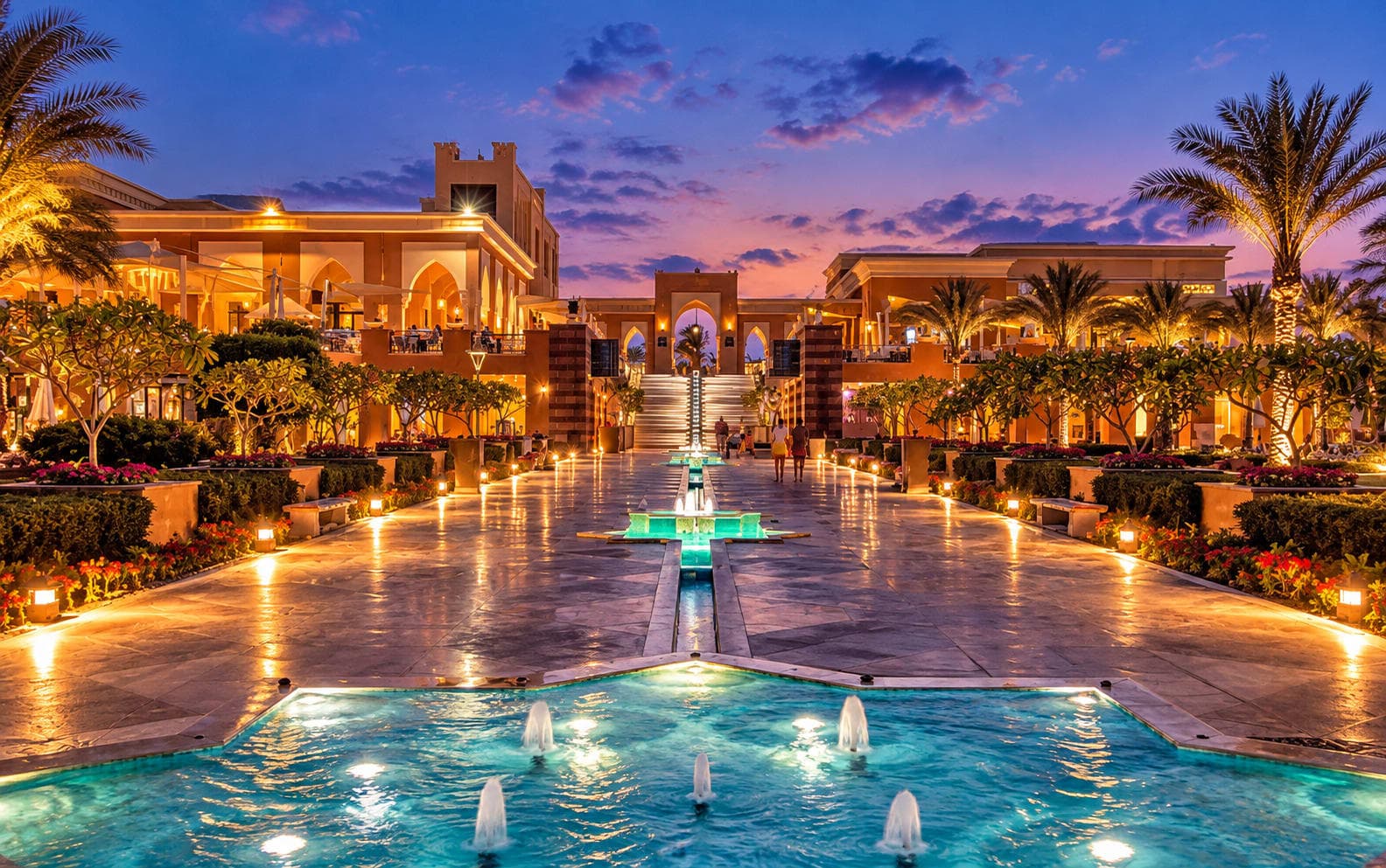 An elegant North Coast resort glowing at sunset with illuminated fountains, palm trees, and luxury architecture.