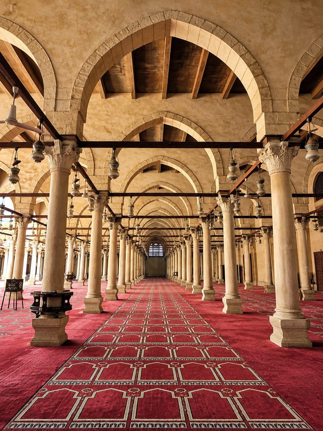 The historic prayer hall of the Mosque of Amr ibn al-As in Cairo, featuring rows of stone columns, Islamic arches, and red prayer carpets.